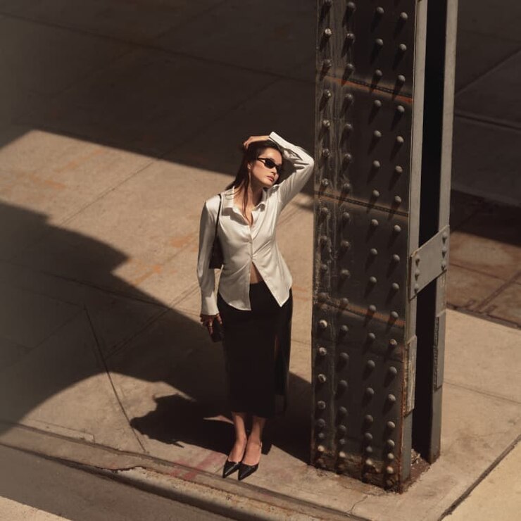 Model is wearing an off white cinched satin blouse and a black midi skirt.